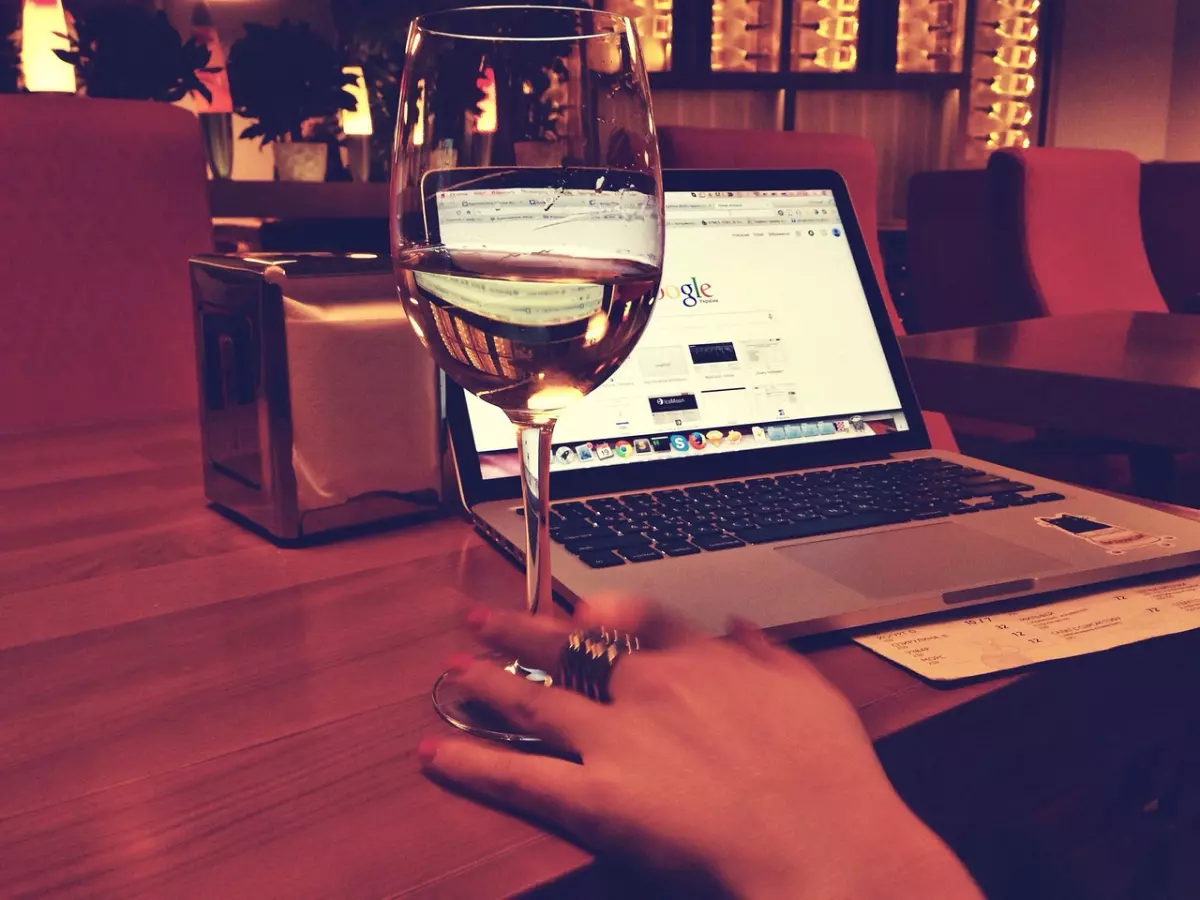 A laptop with Google's logo open on the screen, a glass of white wine, and a woman's hand resting on the table.